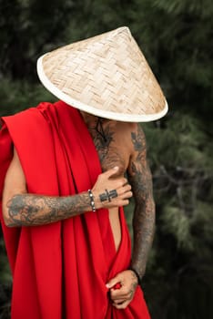 Sexy male monk in a red cape and an Asian triangular hat with tattoos in nature