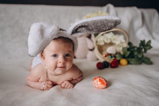 Cute little baby wearing bunny ears for Easter. Near Easter eggs. looks into the camera and smiles. High quality photo