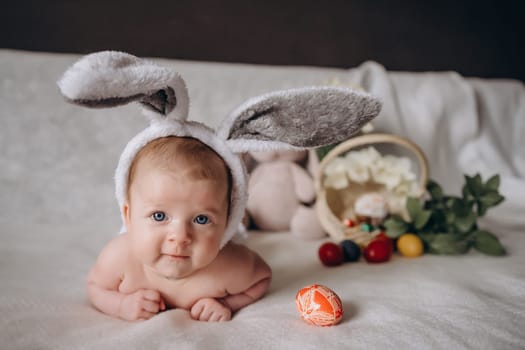 Cute little baby wearing bunny ears for Easter. Near Easter eggs. looks into the camera and smiles. High quality photo