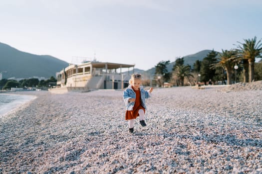 Little girl walks along a pebble beach against the backdrop of green palms and a building. High quality photo