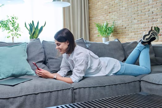 Young smiling happy woman using smartphone, lying on sofa at home in living room. Internet online services, communication, mobile applications, technologies for leisure study work lifestyle