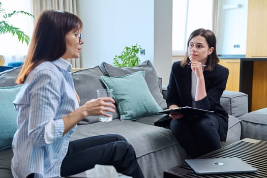 Female psychologist is having therapy session with middle-aged woman patient sitting on sofa in office. Psychology, psychotherapy, therapy, counseling, treatment, mental health concept