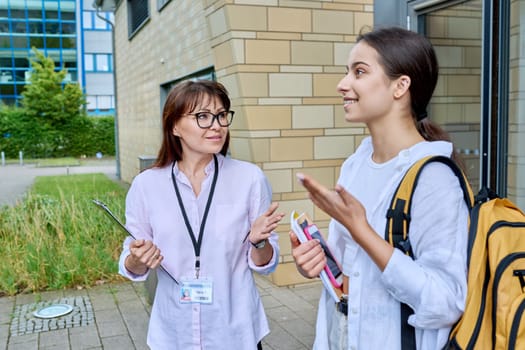 Teenage girl high school student with backpack talking to female teacher, mentor, coach, standing outdoors on educational building background. Adolescence, education, knowledge, communication