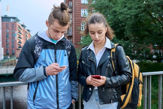 Teen friends guy and girl standing together holding smartphones looking at screen using mobile phones outdoor on city. Internet digital technology applications for leisure study communication