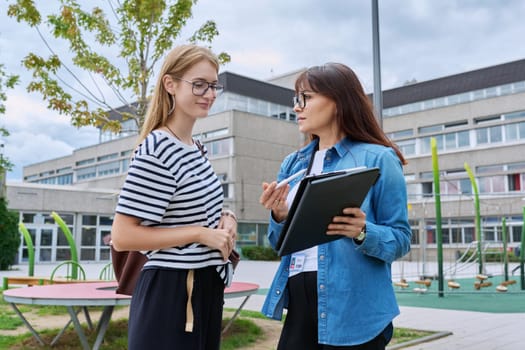 Talking female teacher and teenage high schoolgirl outdoor, school building background. Meeting communication student girl with backpack and mentor counselor. Education, adolescence, learning concept