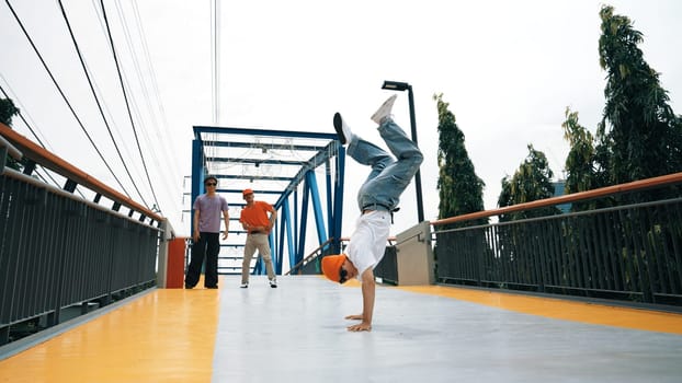 Panorama shot of professional hipster perform b-boy performance and energetic footstep. Skilled happy man practice break dance while friend cheering him at bridge. Outdoor sport 2024. Sprightly.