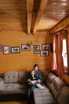 Mom reads a book to a little girl sitting on her lap on the sofa in a wooden house. High quality photo