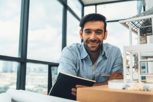 Smiling smart architect engineer looking at camera while sitting near house model. Happy confident businessman wearing casual outfit holding notebook while poses at camera. Civil engineering. Tracery.