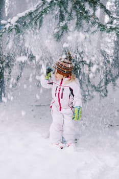 Little girl in a ski suit stands adjusting her hat under snowfall from a Christmas tree in the forest. High quality photo