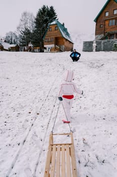 Dad squats on a hill and watches a little girl go up with a sled on a string. Back view. High quality photo