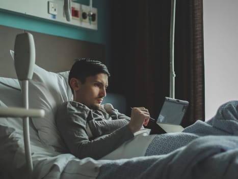 Portrait of one young sad caucasian guy lying in bed with a tablet in a hospital room near the window, close-up view from below.