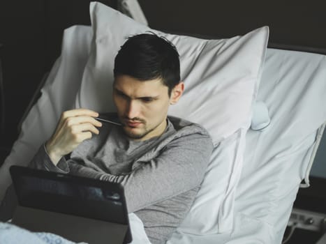Portrait of one young pensive caucasian guy lying in bed with a tablet in a hospital room near the window, top view close-up.