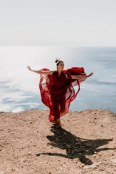 Side view a Young beautiful sensual woman in a red long dress posing on a rock high above the sea during sunrise. Girl on the nature on blue sky background. Fashion photo.