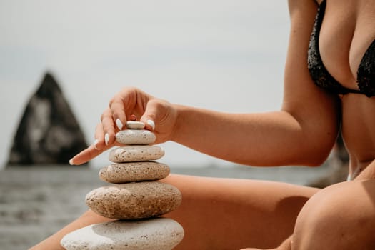 Woman bilds stones pyramid on the seashore on a sunny day on the blue sea background. Happy holidays. Pebble beach, calm sea, travel destination. Concept of happy vacation on the sea, meditation, spa