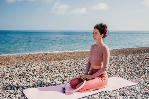 Middle aged well looking woman with black hair doing Pilates with the ring on the yoga mat near the sea on the pebble beach. Female fitness yoga concept. Healthy lifestyle, harmony and meditation.