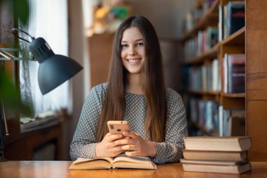 Happy female student with phone reading books in library, looking to the camera