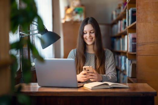 Female student reading books, prepare research as her home assignment using modern technologies: laptop and mobile phone with the Internet