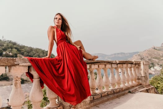 Side view a Young beautiful sensual woman in a red long dress posing on a volcanic rock high above the sea during sunset. Girl on the nature on overcast sky background. Fashion photo