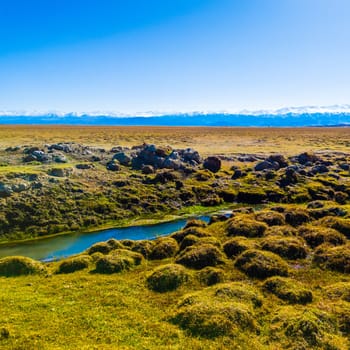 field covered with dry grass bumps and small creek with distant high mountains on the horizon, wide angle surface level view