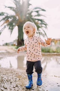 Laughing little girl in rubber boots jumps in a puddle, raising splashes. High quality photo