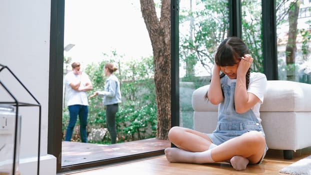 Stressed and unhappy young girl huddle in corner, cover her ears blocking sound of her parent arguing in background. Domestic violence at home and traumatic childhood develop to depression. Synchronos