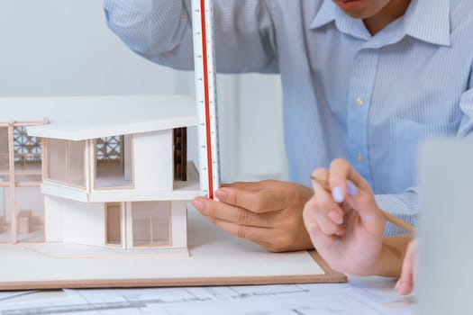 Professional male asian architect using ruler to measure house model length while young beautiful caucasian colleague using laptop to analyzed data on meeting table with house model. Immaculate.