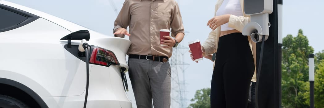 Young couple with coffee recharge EV car battery at charging station connect to electrical industrial power grid. Couple with shopping and travel using eco electric car lifestyle.Panorama Expedient