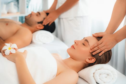 Caucasian couple enjoying relaxing anti-stress head massage and pampering facial beauty skin recreation leisure in dayspa modern light ambient at luxury resort or hotel spa salon. Quiescent