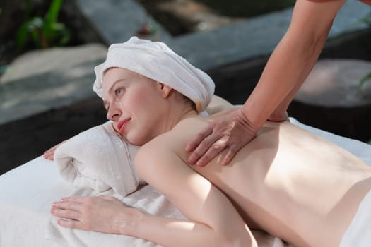 Beautiful young woman received a back massage on a spa bed from professional masseuse. Attractive female relaxes deeply by skilled hands of the massage therapist. Surrounded with nature. Tranquility.