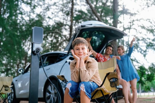 Little boy portrait sitting on camping chair with his family in background. Road trip travel with alternative energy charging station for eco-friendly car concept. Perpetual