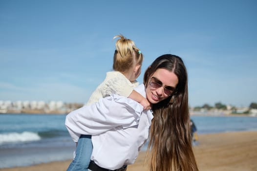 Lifestyle portrait of authentic family of one young mom carrying her cute child girl on her back, enjoying a happy day on the beach. Maternity lifestyle. People. Leisure activity. Weekend activity