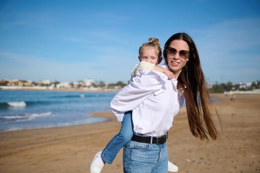 Beautiful and carefree young Caucasian woman, loving mom carrying on shoulders her adorable kid, dreamily looking away, walking together on the sandy beach on sunny warm spring or autumn day