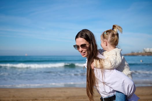 Mom and child girl kid enjoy and fun outdoor lifestyle on the beach. Young delightful Caucasian mother giving piggyback to her lovely daughter, walking along the waves breaking on the Atlantic shore