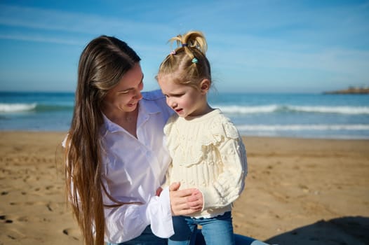 Mother and daughter spending happy time together on the beach on beautiful warm sunny day, sitting on the sandy beach against waves splashing while crashing on the Atlantic shore. Family. Childhood