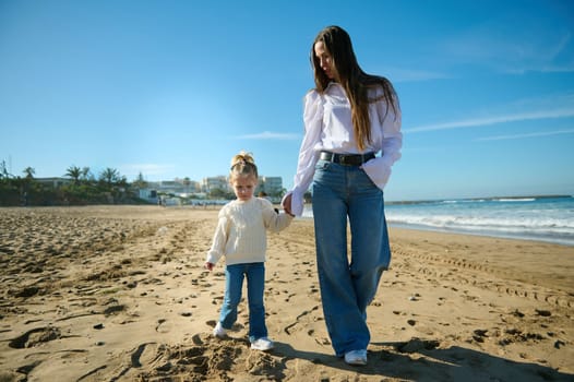 Full length portrait of a happy woman, carefree happy mother holding the hands of her lovely child girl, walking together on the sandy beach on a warm sunny day. People. Lifestyle. Leisure activity