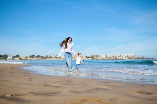 Happy Caucasian young mother and her adorable daughter girl, running barefoot together on sandy beach in sea water in summer in warm ocean waves. Full length portrait. Family pastime. Togetherness