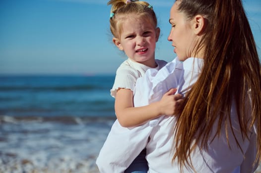 Adorable blonde little baby girl looking at the camera, sitting on the arms of her mother carrying her while walking along the Atlantic ocean beach. Mom and daughter together. Happy carefree childhood