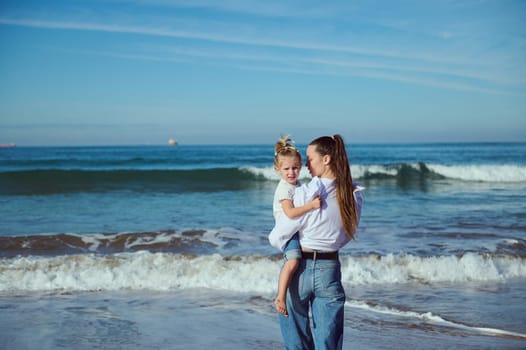 A beautiful young mother in white casual shirt and blue jeans, carries her baby in her arms, an adorable little girl, walking together barefoot on the beach. Mom and daughter. People and nature
