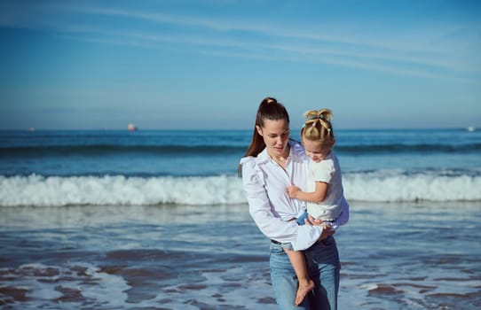 Beautiful Caucasian young woman talking with her daughter while carrying her, walking together on the beach. Maternity leave lifestyle. Family relationships. Happy carefree childhood. Copy ad space