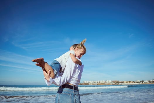 Authentic portrait of Caucasian pretty happy young woman, mother playing with her cute baby girl on the beach, turning around, laughing, walking leaving footstep on the wet sand. Family relationships