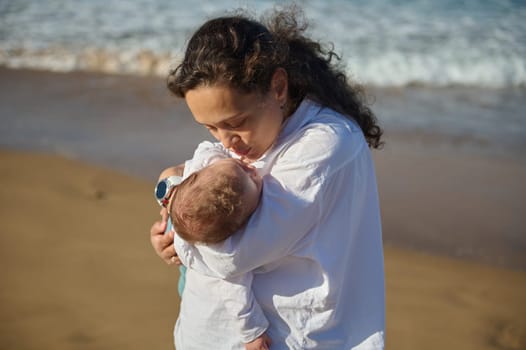 Loving affectionate young mother kissing her baby boy, holding him in her arms while walking along a sandy beach on warm sunny day. Mom and son. Tenderness. Love. Care. Affection. Babyhood. Maternity