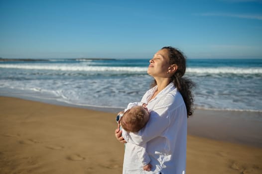 Delightful curly haired multi ethnic young woman, a loving mother carrying her adorable baby boy, standing with her eyes closed on the sandy beach on warm sunny day, enjoying happy time together