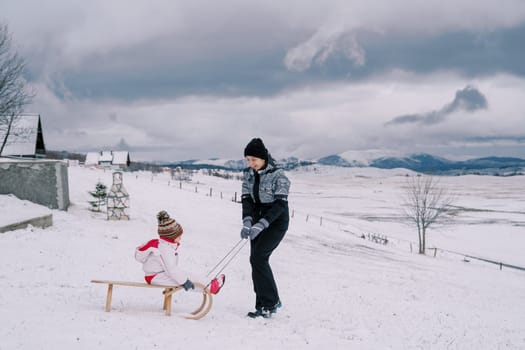 Smiling mother carries little girl on wooden sled along snowy hill. High quality photo