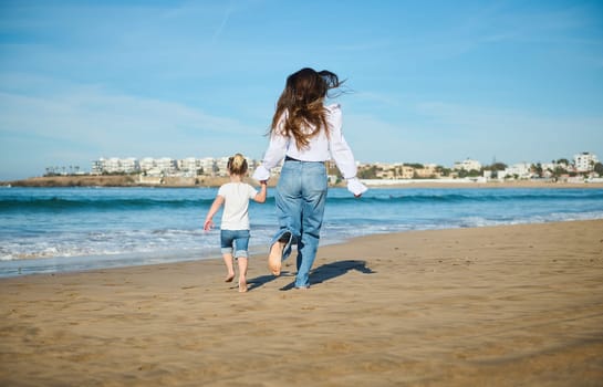 Rear view of a young mother and daughter holding hands, running barefoot on the wet sand, leaving footsteps, enjoying happy moments together on beautiful sunny day. People and lifestyle
