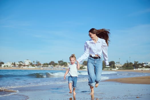 Authentic portrait of Caucasian happy mom and daughter smile while run along the Atlantic shore, enjoying beautiful waves washing their feet while pounding on the shore, leaving footsteps on the sand