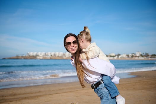 Happy Caucasian family of a young loving caring mother carrying little daughter on her back and walking on tropical sandy beach. Mom and child girl kid enjoy and fun outdoor lifestyle on the beach