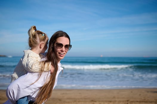 Young woman, happy mother carrying her adorable little kid girl, lovely daughter on her back, giving her a piggyback ride while walking together on the beach. Beautiful waves breaking on the shore
