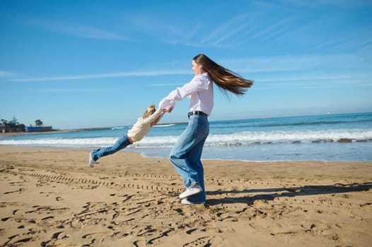 Full length portrait of a happy young woman, loving mother spinning her little child girl, an adorable daughter while playing on the sandy beach. The concept of family pastime and relationship