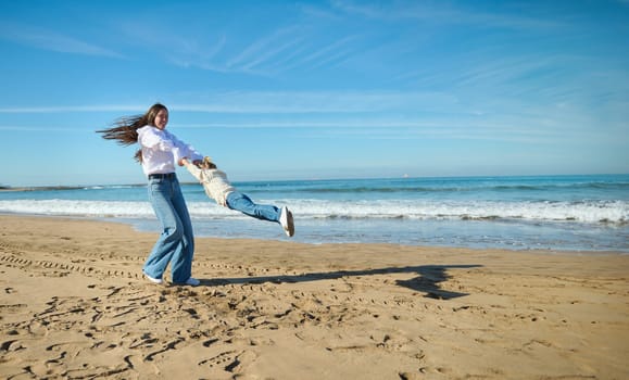 Mother and child daughter having fun together on the beach. Loving mom spinning her cute kid girl around on the sandy beach. People. Active healthy lifestyle. Leisure activity