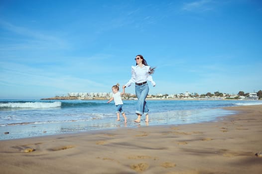 Caucasian young mother and daughter holding hands, running barefoot on the wet sand, leaving footsteps, enjoying happy moments together on beautiful sunny day. People and lifestyle